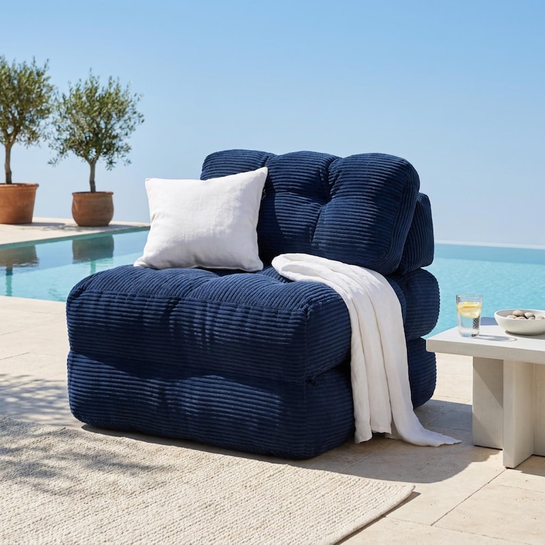 May include: A navy blue corduroy outdoor lounge chair with a white pillow and blanket. The chair is near a pool with olive trees in the background. A small white side table holds a glass of water and a bowl.