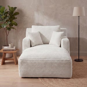 May include: A white corduroy chaise lounge with matching pillows. The lounge has a wide seat and a backrest with two armrests. Two smaller square pillows and a rectangular pillow are on the backrest. A floor lamp and a small wooden side table are in the background.