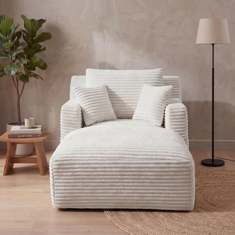 May include: A white corduroy chaise lounge with matching pillows. The lounge has a wide seat and a backrest with two armrests. Two smaller square pillows and a rectangular pillow are on the backrest. A floor lamp and a small wooden side table are in the background.