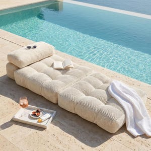 May include: A beige corduroy outdoor lounge chair with a matching headrest, a white tray with a drink, figs, and sunscreen, and a white waffle-weave robe. Sunglasses and a book rest on the chair near a pool.