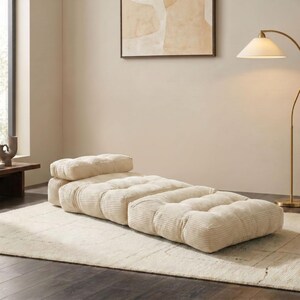 May include: A cream-colored, corduroy-style floor lounger with a rectangular shape and a separate headrest. The lounger is placed on a cream-colored rug in a well-lit room. The design is modern and minimalist.