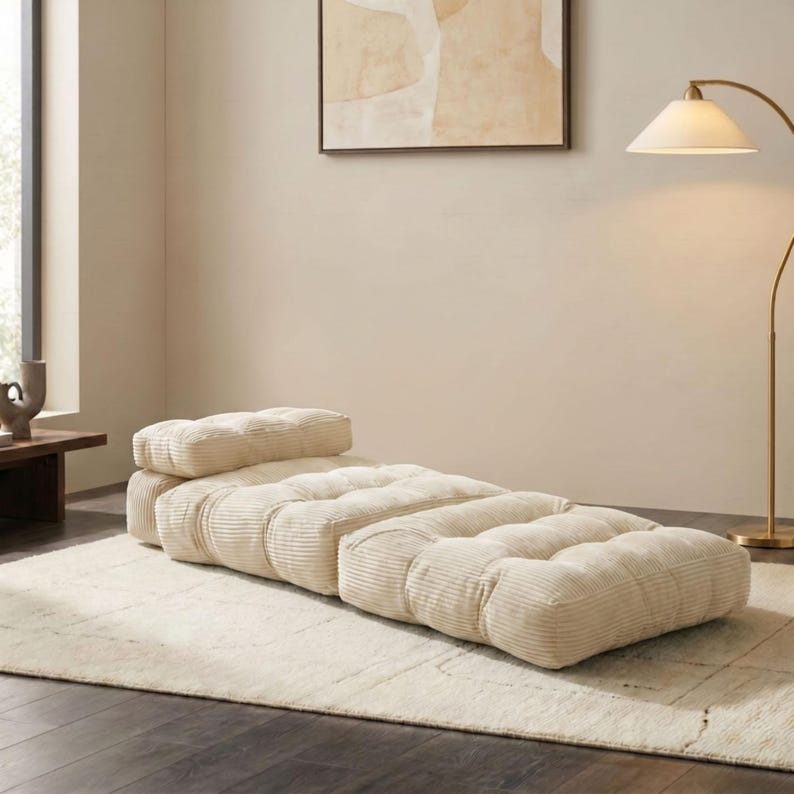 May include: A cream-colored, corduroy-style floor lounger with a rectangular shape and a separate headrest. The lounger is placed on a cream-colored rug in a well-lit room. The design is modern and minimalist.