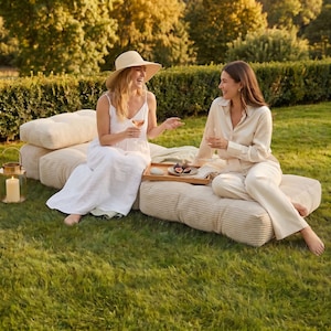 May include: Two women enjoying a picnic outdoors. They are seated on beige corduroy cushions on a grassy lawn. A wooden tray holds figs and a drink. One woman wears a white dress and hat, the other a cream-colored outfit.