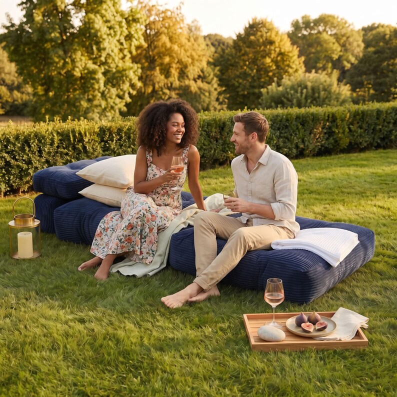 May include: A couple enjoys a relaxing outdoor setting on navy blue corduroy floor cushions. A wooden tray holds figs and a glass of wine. A gold lantern with a candle sits nearby. Beige pillows and a white blanket add comfort.