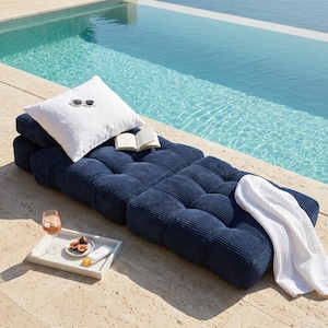 May include: A navy blue corduroy lounge chair with a white pillow featuring sunglasses embroidery. A white waffle-weave blanket rests on the chair. A tray with a drink, figs, and a book are nearby, next to a pool.