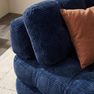 May include: Close-up of a section of a blue corduroy sofa with a square-stitched design. A rust-colored pillow rests against the back cushions. The sofa has a soft, inviting appearance, perfect for a living room or den.