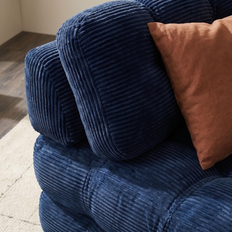 May include: Close-up of a section of a blue corduroy sofa with a square-stitched design. A rust-colored pillow rests against the back cushions. The sofa has a soft, inviting appearance, perfect for a living room or den.