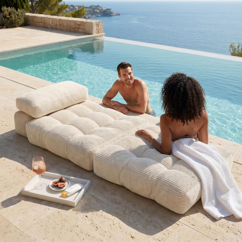 May include: A cream-colored, modular outdoor lounge set with a tufted design. The set includes a backrest and multiple seating sections. A white tray with a drink, figs, and sunscreen sits nearby. A person is in the pool, and another is lounging on the set.