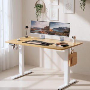Può includere: Una scrivania regolabile in altezza in tonalità legno chiaro con gambe bianche. La scrivania ha due monitor per computer, una tastiera, un mouse e un tappetino nero. Un paio di cuffie sono appese al lato. Sul tavolo ci sono anche una piccola lampada e un portapenne.