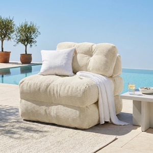 May include: A cream-colored corduroy armchair with a white pillow and a white blanket draped over it. The chair is on a patio with a pool and trees in the background. A small white table with a glass of water and a bowl is next to the chair.