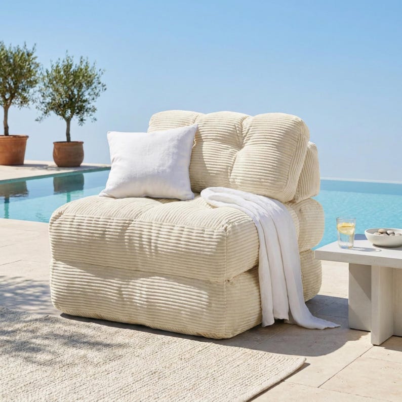 May include: A cream-colored corduroy armchair with a white pillow and a white blanket draped over it. The chair is on a patio with a pool and trees in the background. A small white table with a glass of water and a bowl is next to the chair.
