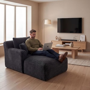 May include: A dark gray corduroy sectional sofa with an attached ottoman. The sofa has a high back and armrest, and a person is sitting on it with a laptop. The room has a neutral color palette with a wooden floor and a large window.