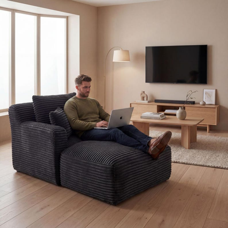 May include: A dark gray corduroy sectional sofa with an attached ottoman. The sofa has a high back and armrest, and a person is sitting on it with a laptop. The room has a neutral color palette with a wooden floor and a large window.