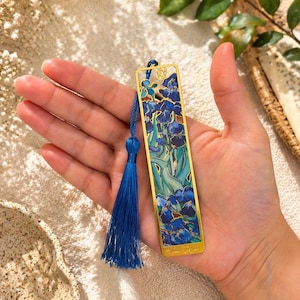 May include: A gold-toned metal bookmark featuring a blue floral design with a matching blue tassel. The bookmark has a rectangular shape with a detailed illustration of blue irises and green leaves. The tassel is attached to the top of the bookmark.