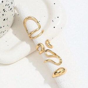 Könnte beinhalten: Ein Set aus fünf goldfarbenen Ringen mit unregelmäßigen, organischen Formen. Die Kollektion umfasst einen großen offenen Ring, ein kleines geschwungenes Teil, ein gewelltes Band, einen glatten abgerundeten Ring und einen kleinen halbmondförmigen Ring.