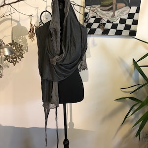 May include: A dark gray scarf with a hood is draped over a black mannequin. The scarf has a flowing, layered design with a textured appearance. The mannequin is on a black stand, and the background includes artwork and decorative elements.