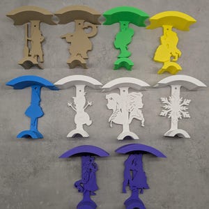 Peut inclure: Une collection de figurines décoratives en forme de parapluie colorées. Chaque figurine présente un design de silhouette unique, comprenant des personnages et des formes. La palette de couleurs comprend le beige, le vert, le jaune, le bleu, le blanc et le violet. Les figurines sont disposées sur une surface grise.