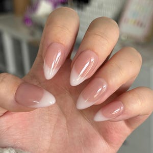 Puede incluir: Una mano que muestra uñas artificiales en forma de almendra. Las uñas presentan una base rosa natural con un diseño de punta francesa blanca. Las uñas son brillantes y están bien cuidadas, mostrando un estilo clásico y elegante.