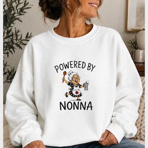 May include: A white crewneck sweatshirt featuring a graphic print of an elderly woman with glasses, wearing a black and white polka dot dress and apron, holding a wooden spoon and a moka pot, with the text "POWERED BY" above and "NONNA" below.