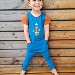 Könnte beinhalten: Blauer Overall mit orangefarbenen Ärmeln und einem Raketenmotiv. Der Overall hat zwei Vordertaschen und wird von einem Kind getragen. Der Hintergrund ist eine Holzwand.