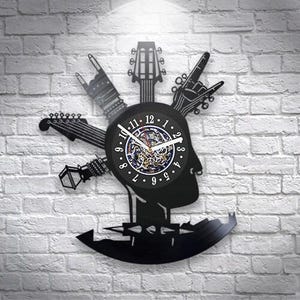 Puede incluir: Un reloj de pared negro hecho de un disco de vinilo, con la silueta de una cabeza con una cresta hecha de mástiles de guitarra y gestos con las manos. El dial central muestra números blancos del 1 al 12 y manecillas blancas sobre engranajes.