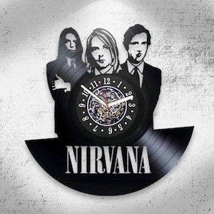 Può includere: Un orologio da parete realizzato con un disco in vinile nero con una sagoma ritagliata della band Nirvana. Il quadrante mostra numeri bianchi da 1 a 12, lancette bianche e un meccanismo a ingranaggi visibile. La parola NIRVANA è stampata in bianco.