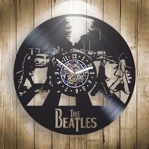 Pode incluir: Um relógio de parede circular feito de um disco de vinil preto com uma silhueta cortada a laser dos Beatles atravessando uma faixa de pedestres. O centro exibe um mostrador com números e ponteiros brancos, com o texto The Beatles na parte inferior.