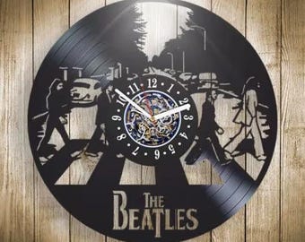 Puede incluir: Un reloj de pared circular hecho de un disco de vinilo negro con una silueta cortada con láser de The Beatles cruzando un paso de peatones. El centro muestra una esfera con números y manecillas blancas, con el texto The Beatles en la parte inferior.