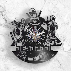 Può includere: Un orologio da parete circolare realizzato con un disco in vinile nero che presenta diverse illustrazioni ritagliate del personaggio Deadpool in varie pose. Il quadrante centrale mostra numeri bianchi da 1 a 12 con lancette bianche.