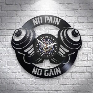 Puede incluir: Un reloj de pared circular hecho de un disco de vinilo negro con dos mancuernas y el texto NO PAIN NO GAIN. La esfera central muestra números blancos del 1 al 12 con manecillas blancas sobre un diseño mecánico detallado de engranajes.