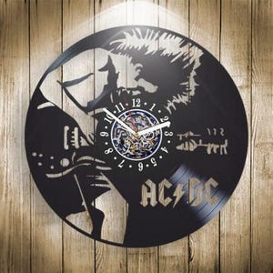 Puede incluir: Un reloj de pared circular hecho de un disco de vinilo negro con la silueta de un guitarrista cortada con láser. El centro muestra una esfera blanca con números del uno al doce y manecillas blancas. El texto AC/DC está cortado abajo a la derecha.