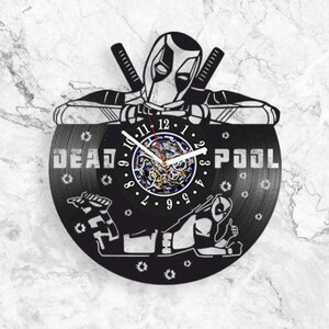 Può includere: Un orologio da parete circolare realizzato con un disco in vinile nero con un design ritagliato del personaggio Deadpool. Il quadrante mostra numeri bianchi, lancette bianche e il testo DEAD POOL. Dettagli di ingranaggi sono visibili al centro.