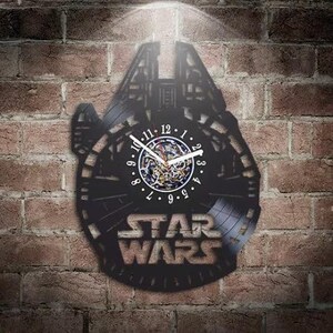 May include: A black wall clock crafted from a vinyl record, shaped like the Millennium Falcon spaceship. The center features a white clock face with black numbers and hands, and the text STAR WARS is cut out at the bottom of the record.