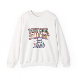 Puede incluir: Sudadera blanca con el texto "Barely Coping, Still Iconic" en una fuente retro colorida. El gráfico presenta un personaje de dibujos animados en un sofá sosteniendo una taza. La sudadera es de estilo casual y cómodo.