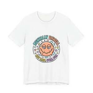 Puede incluir: Camiseta blanca con una ilustración de dibujos animados de una cara sonriente de color melocotón. El diseño incluye las palabras "Mentally Unwell But Like, Still Fun" en un diseño circular, con estrellas, corazones y otras formas pequeñas.