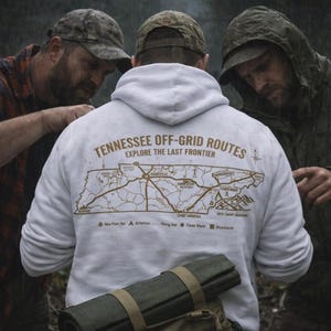 May include: Three people in outdoor gear examine a white hoodie with a map graphic. The hoodie features the text "Tennessee Off-Grid Routes" and "Explore The Last Frontier." A rolled-up sleeping pad rests on a backpack.