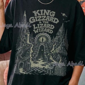 Könnte beinhalten: Schwarzes T-Shirt mit dem Text "King Gizzard & The Lizard Wizard" und einer detaillierten Illustration. Die Person trägt das Shirt in einem Raum mit Postern und Schallplatten, was eine Retro-Atmosphäre erzeugt.
