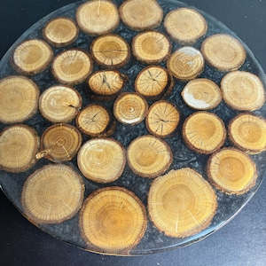 May include: A round decorative piece featuring numerous small, light brown wood slices embedded in a dark gray resin. The wood slices have visible growth rings and are arranged in a circular pattern, creating a natural, rustic aesthetic.