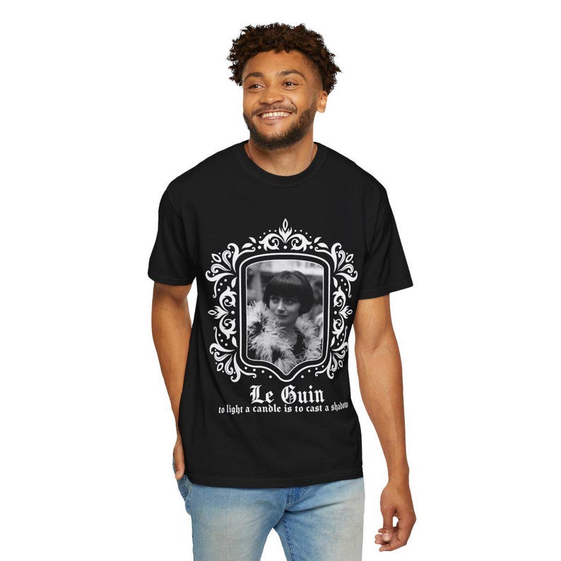 May include: Black t-shirt featuring a black and white portrait within a decorative white frame. The portrait is of a woman with short hair and a feathered boa. The text "Le Guin" and a quote are below the portrait.