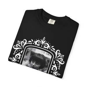 May include: Black t-shirt featuring a black and white framed portrait of a person with short hair. The frame is white with ornate floral and scrollwork designs. The shirt has a crew neck and a small tag.