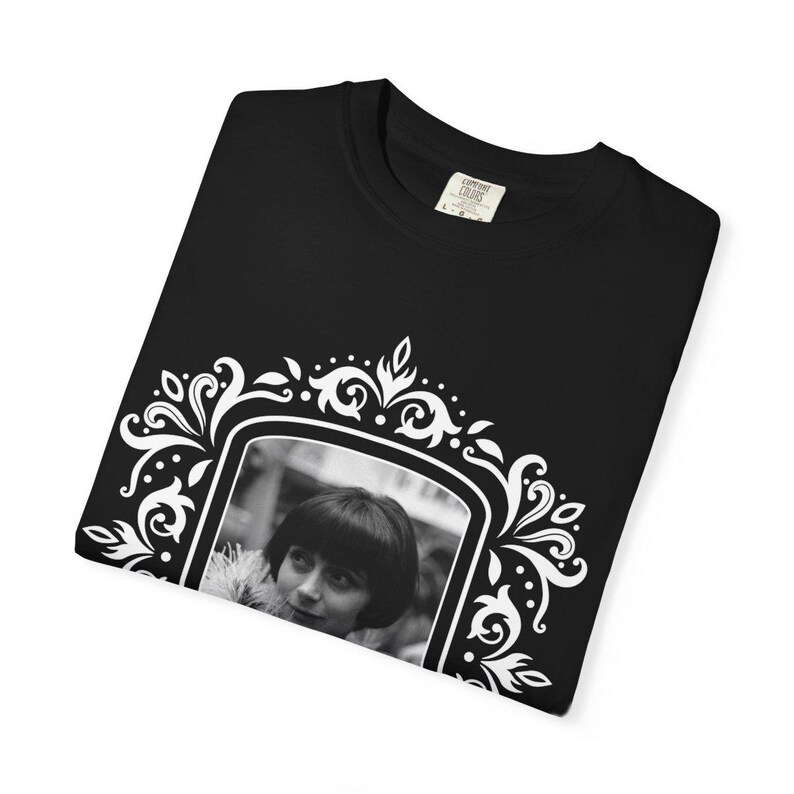 May include: Black t-shirt featuring a black and white framed portrait of a person with short hair. The frame is white with ornate floral and scrollwork designs. The shirt has a crew neck and a small tag.