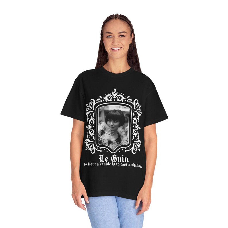 May include: Black t-shirt featuring a black and white portrait within a decorative white frame. The portrait is of a woman with short hair and a feathered boa. The text "Le Guin" and a quote are printed below the portrait.