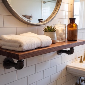 May include: A dark wood floating shelf mounted with black industrial pipe brackets. The shelf holds a folded white towel, a small potted succulent, a clear glass jar filled with white cotton balls, and a tall amber glass bottle with a cork stopper.