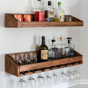 May include: Two wooden bar shelves mounted on a white wall. The top shelf holds glasses and liquor bottles, including Jack Daniel's. The bottom shelf has a rack for hanging wine glasses and more bottles.
