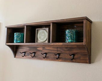 May include: A dark stained wooden wall shelf with three compartments and five black hooks. The shelf holds two teal glass candle holders and a framed sign that reads "Live Simply".