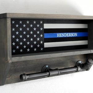 May include: A gray wooden wall shelf with a thin black shelf and a blue and white American flag with a thin blue line and the name "Henderson" printed on it. The shelf has two black metal pipe hooks.