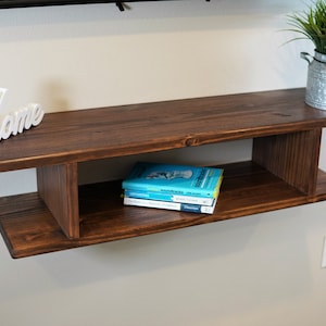 May include: A dark brown wooden floating shelf with two shelves and a decorative white "Home" sign on the top shelf. There are three books on the lower shelf, and a small galvanized metal planter with a yellow sunflower on the top shelf.