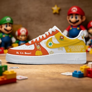 May include: White, orange, and yellow custom sneakers featuring Princess Daisy from the Super Mario franchise. The shoe has the text "Hi, I'm Daisy!" and "Princess Daisy" printed on it. The sneakers are on a wooden surface.