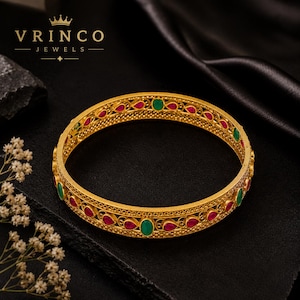 May include: A gold bangle bracelet featuring an intricate filigree pattern with alternating teardrop-shaped red stones and oval-shaped green stones. The metal surface has a textured finish, and the brand name VRINCO JEWELS is printed in the top corner.
