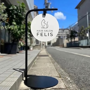 May include: A black metal sign for a hair salon, featuring a white circular sign with a cat silhouette and the text "HAIR SALON FELIS". The sign is mounted on a curved pole with a round base, positioned on a sidewalk.